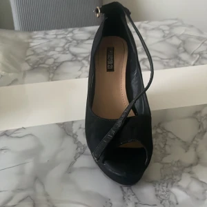 Svarta wedges med öppen tå från Forever 21 - Snygga svarta wedges från Forever 21 med öppen tå och rem runt vristen med guldfärgat spänne. Skorna har hög kilklack och är gjorda i mockaimitation. Perfekta för dig som vill ha både höjd och stabilitet.