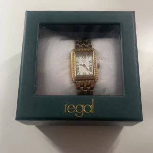 Guldig rektangulär klocka från Regal - Stilren klocka från Regal med rektangulärt ur i guldton och vit urtavla med romerska siffror. Länken är i metall och har en klassisk design. Runt urtavlan sitter små gnistrande stenar som ger extra bling. Perfekt accessoar för dig som gillar en elegant look.