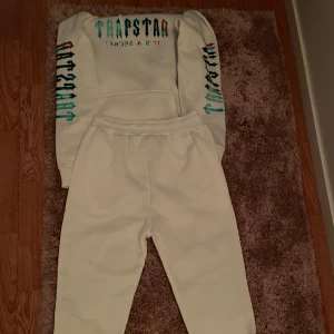 Vita mjukisbyxor från Trapstar och hoodie  - Säljer ett par vita mjukisbyxor från Trapstar med elastisk midja och tryckt logga på ena benet. Byxorna är i mjukt bomullsmaterial och har en relaxed fit som är perfekt för chill dagar. Snygg och enkel design med streetkänsla. Med det kan du också få en hoodie som också är från Trapstar med loggan på armarna och på bröst höjd med Texten trapstar.