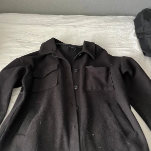 Svart Kappa i ull med fickor - Säljer en svart kappa i ull med klassisk krage och stora knappar framtill. Jackan har två bröstfickor och två sidofickor, perfekt för en chill och stilren look. Passar dig som gillar minimalistiska och tidlösa plagg.