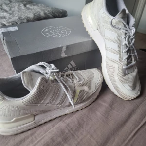 Vita Adidas ZX 750 sneakers - Snygga Adidas ZX 750 sneakers i vitt med mesh och syntetmaterial, klassiska tre ränder på sidan och coola detaljer i ljusgrönt baktill. Skorna har snörning och en bekväm platt sula, perfekta för dig som gillar sportig stil. Använd ett par gånger
