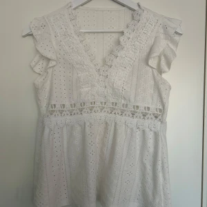 Vit spetsblus med volang och broderi - Superfin vit blus med v-ringning, spetsdetaljer och broderade mönster. Ärmlös modell med volang vid axlarna och dekorativt hålmönster. Står tyvärr inte vilken storlek men skulle säga att det är en xs, kan nog funka på s också!!