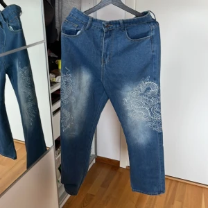 Blå jeans med drakbroderi från BH - Snygga blå jeans från BH i storlek L  men passar M med kinesisk vitt drakbroderi på benen. Jeansen har raka ben, klassisk femficksdesign och coola slitningar framtill. 