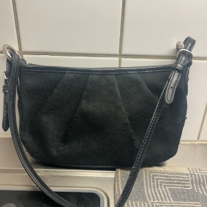 Svart axelväska från DKNY - Snygg svart axelväska från DKNY med diskret logomönster i tyget. Väskan har justerbar axelrem i skinn och silverfärgade metalldetaljer. Perfekt storlek för det viktigaste och stilren design som funkar till många outfits.
