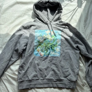 Grå hoodie med tryck - Grå hoodie från Monki i storlek S med en stor färgglad blommig print framtill. Tröjan har huva med snörning och är tillverkad i mjuk bomullsmix. 