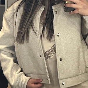 Beige bomberjacka från Zara - Snygg beige bomberjacka från Zara med ribbade muddar och krage. Jackan har tryckknappar framtill, två fickor och en loose passform. Perfekt för dig som gillar en clean och stilren look. Materialet känns mjukt och är troligtvis en blandning av ull och syntet.
