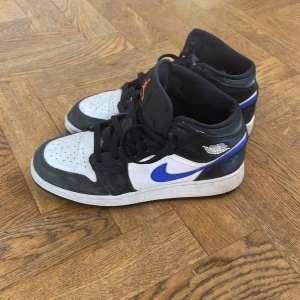 Nike Air Jordan 1 svart/blå sneakers - Säljer ett par Nike Air Jordan 1 sneakers i svart, vitt och blått med klassisk blå swoosh och orange Jumpman-logga på plösen. Dem är i färgkombination racer blue och Skorna har snörning, rund tå och hög siluett. Perfekta för dig som vill köpa ett par stilrena sneakers för sommaren ☀️🔥men vill inte spendera för mycket. Säljer till ett väldigt bra pris💯💯