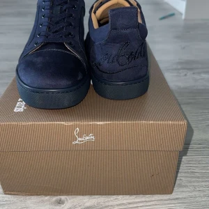 Mörkblå sneakers från Louboutin - Snygga mörkblå sneakers från Christian Louboutin i mocka med klassisk röd sula och diskret broderad logga på hälen. Skorna har rund tå, snörning och en stilren design som sticker ut med den ikoniska röda undersidan.