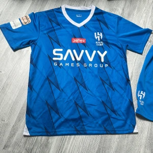 Al Hilal Neymar Jr #10 matchtröja set -  Mycket bra skick på tröjan förutom shortsen som har en fläck på sig,Säljer ett blått Al Hilal matchställ med Neymar Jr #10. Tröjan har korta ärmar, V-ringad vit krage och svart blixtmönster. Tryck med SAVVY Games Group och Jahez på framsidan. Shorts med nummer 10 och klubbmärke. Materialet är lätt och andas, perfekt för fotboll.