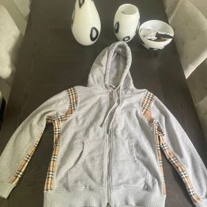 Burberry hoodie - Snygg grå hoodie från Burberry med klassiska beige-rutiga paneler längs ärmarna och axlarna. Tröjan har huva med snörning, dragkedja framtill och två fickor. Perfekt för dig som vill ha en stilren men ändå unik look. Passar perfekt om du har storlek M.