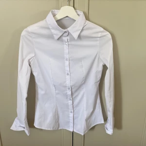 Vit skjorta från Angel Swingz - Klassisk vit skjorta från Angel Swingz med lång ärm och figurnära passform. Skjortan har krage, knäppning framtill med vita knappar och är tillverkad i ett mjukt bomullstyg. Stretchigt material. Perfekt för en clean och stilren look. Passar en lite S eller XS.