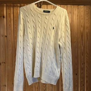 Vit kabelstickad tröja Polo Ralph Lauren - Kabelstickad vit tröja från Polo Ralph Lauren med klassisk rund halsringning och broderad marinblå logga på bröstet. Tröjan är i bomull och har ribbade muddar vid ärmslut och nederkant. Perfekt för lager-på-lager under hösten och vintern.