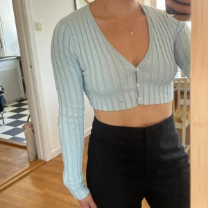 Ljusblå croppad kofta från Monki - Superfin ljusblå croppad kofta från Monki med ribbat tyg och v-ringning. Tröjan har långa ärmar och knäpps med tre knappar framtill. Perfekt att styla med höga byxor för en trendig look.