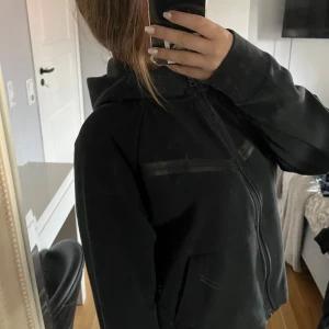 Svart Nike Tech - Svart hoodiejacka från Nike med dragkedja framtill och huva. Jackan har diskreta svarta detaljer över bröstet och två fickor. Perfekt för chill dagar eller när du vill ha en sportig look. Det är barnstorlek, men om man vanligtvis är S så sitter den perfekt