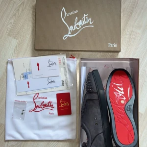 Christian Louboutin grå mocka sneakers - Snygga grå sneakers från Christian Louboutin i mocka med ikoniska röda sulor och coola nitar på tån. Skorna har snörning och en rund tå, perfekt för dig som vill sticka ut med exklusiv streetstyle.