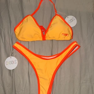 Gul och orange bikini från Boutine LA x Chooks - Superfräsch bikini från Boutine Los Angeles x Chooks i en snygg gul färg med orangea kanter. Klassisk triangelmodell upptill med knyt i nacken och smala axelband. Underdelen har högt skurna ben. Perfekt för stranden eller poolen! Båda delarna är i strl L men är små i storleken. Inköpspris 900kr