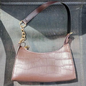 Brun axelväska med krokoprägling - Säljer en brun axelväska i syntetmaterial med krokodilpräglad yta och guldfärgade metalldetaljer. Väskan har en kort axelrem och dragkedja upptill. Perfekt för dig som vill ha en trendig och stilren accessoar till din outfit.