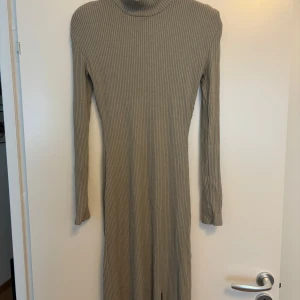 Ribbad beige långklänning Gina Tricot - Supersnygg ribbad långklänning i beige/grön från Gina Tricot. Klänningen har lång ärm, hög hals och en slits framtill som ger en cool vibe. Figurnära passform och stretchigt material som sitter perfekt. Perfekt för dig som gillar stilrena och trendiga plagg.