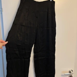 Svarta vida byxor från Zara - Snygga svarta byxor från Zara med hög midja och vida ben. Byxorna har en lätt glansig finish och stängs med dragkedja och knapp framtill. Perfekta för en stilren look och passar till många olika outfits.