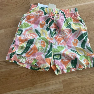 Färgglada linneshorts med snörning - Snygga linneshorts från Disa i storlek 36 med tropiskt mönster i rosa, grön, orange, gul och vit. Shortsen har elastisk midja och snörning för extra komfort. Perfekta för varma sommardagar när du vill sticka ut med färg och stil.