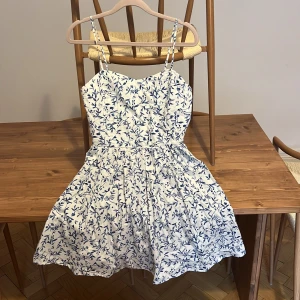 Vit och blå blommig sommarklänning H&M - Supersöt kort klänning från H&M i vitt med blått blommönster. Smala justerbara axelband, smockad rygg och utställd kjol. Perfekt för varma dagar och ger en fräsch och ungdomlig vibe. Gjord i mjuk bomull som andas.