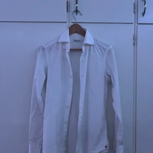 Vit skjorta från Loro Piana, stl 38 - Klassisk vit skjorta från Loro Piana i 100% bomull. Skjortan har lång ärm, knappar framtill och en snygg krage. Italiensk design med diskret logga på manschetten. Perfekt för dig som gillar stilrena och exklusiva plagg.