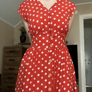 Röd prickig långklänning med knappar - Superfin röd klänning med vita prickar och knappar framtill. Klänningen har v-ringning, markerad midja med knytband bak och är ärmlös. Längden är mellanlång och tyget är lätt och skönt, perfekt för varma dagar. Super chic och retro, påminner om 50/60-talet.