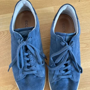 Blå mockasneakers från Ecco strl 42 - Snygga blå sneakers från Ecco i mocka med vita detaljer och klassisk snörning. Skorna har en rund tå, platt sula och innersula i vegetabiliskt garvat skinn. Perfekta för dig som gillar stilrena och bekväma sneakers med en modern touch.