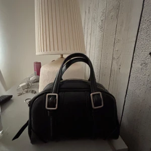 Svart handväska med silverdetaljer - Snygg svart handväska i skinn med två handtag och coola silverfärgade spännen framtill. Väskan har dragkedja upptill och en stilren, rundad form som passar perfekt till din vardagsstil. Zara