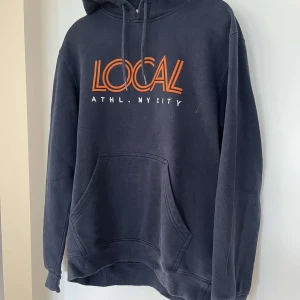 Svart hoodie LOCAL från H&M - Svart hoodie från H&M med orange och vit text 'LOCAL ATHL. NYCITY' på bröstet. Klassisk känguruficka framtill, huva med snörning och mjukt material på insidan. Perfekt för chill dagar eller när du vill ha en avslappnad stil.