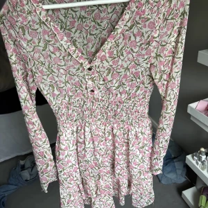Blommig kortklänning med volang - Supersöt kortklänning med rosa och grönt blommönster på ljus botten. Klänningen har v-ringning, smockad midja, långa ärmar och volangig kjol. Perfekt för vår och sommar, ger en romantisk och fräsch vibe.