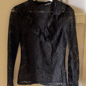 Svart spetsblus med volangdetalj - Supersnygg svart blus i transparent spets med blommigt mönster och volang längs ringningen. Långärmad och figurnära passform, perfekt att styla med jeans eller kjol för en trendig look.