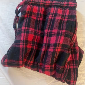 Rödrutiga mjukisbyxor med snörning - Mysiga mjukisbyxor i röd och svart rutig design. Byxorna har resår i midjan och snörning för extra komfort. Perfekta för chill hemma eller när du vill vara extra bekväm. Tillverkade i mjukt bomullstyg.