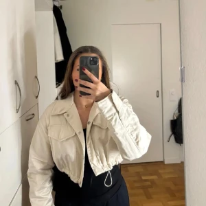 Beige croppad jacka med snörning - Snygg beige croppad jacka med långa ärmar och knappar samt två fickor framtill. Jackan är från Zara och är beige/cremefärgad. Den går att spänna till längst ner beroende på hur tajt/puffig du vill att den ska sitta. Använd fåtal gånger!