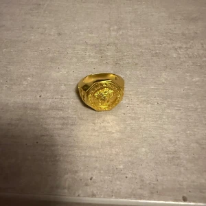 Guldfärgad ring med medusa  - guldfärgad ring med ett detaljerat medusa på ovansidan och mönstrad kant. Ringen har en kraftig design och sticker verkligen ut. Perfekt accessoar för dig som vill ha något unikt och iögonfallande.