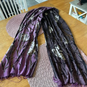 Lila blommig halsduk med fransar - Snygg lila halsduk med stora blommor i vitt och grått samt svarta detaljer. Tunt och mjukt tyg med fransar i ändarna, perfekt att slänga över axlarna för en cool look. Blommönstret ger en unik och trendig vibe.