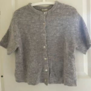 Ljusgrå stickad kofta från Gina Tricot - Supermjuk ljusgrå stickad kofta från Gina Tricot i storlek XS. Koftan har korta ärmar, rund halsringning och knäpps med pärlemorskimrande knappar framtill. Perfekt att slänga över en topp för en chill och stilren look.