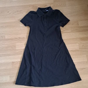 Svart pikéklänning - Svart kortklänning från Divided med klassisk pikékrage och knappar framtill. Klänningen har korta ärmar och lätt utställd passform. Perfekt för en clean och enkel stil. Tillverkad i mjukt material som känns skönt mot huden.💕
