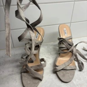 Grå mockasandaler med snörning Vices - Snygga sandaler i grå mocka från Vices med höga klackar och långa snören att vira runt benet. Skorna har öppen tå och breda remmar som ger en trendig look. Perfekta för dig som vill sticka ut med en cool och modern stil.