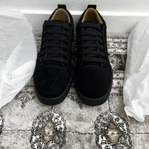 Louboutin skor - Snygga svarta sneakers från Louboutin i mjuk mocka med klassisk röd sula och broderad Louboutin-logga på hälen. Skorna har rund tå, svarta skosnören och en stilren design som sticker ut med den ikoniska röda undersidan.