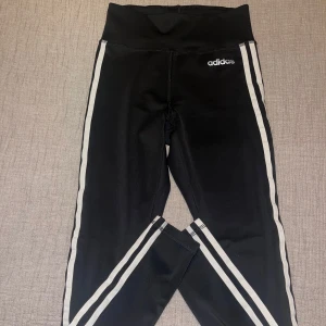 Svarta Adidas leggings - Svarta leggings från Adidas i storlek XS med klassiska vita ränder längs sidorna och Adidas-logga på höften. Hög midja och stretchigt material som sitter tight. Perfekta för träning eller chill. Climalite-material som andas.