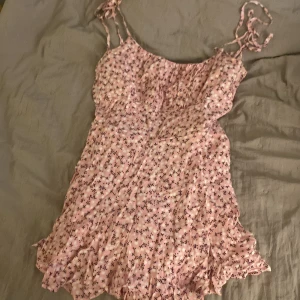Rosa blommig kortklänning FB Sister XS - Supersöt rosa kortklänning (eventuellt byxdress??) från FB Sister i storlek XS. Klänningen har smala knytband, volangkant nertill och ett blommigt mönster i vitt och svart. Lätt och luftig med innerfoder, perfekt för varma dagar. Sitter snyggt och känns riktigt somrig!