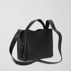 Svart axelväska i skinnlook - Minimalistisk svart axelväska med stilren design och bred axelrem. Väskan har en boxig form och är tillverkad i slätt material med metallnitar på sidorna. Perfekt för dig som gillar enkel och clean stil.