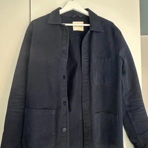 Mörkblå overshirt från A Day's March - Snygg mörkblå overshirt från A Day's March. Jackan har klassisk krage, knäppning framtill och tre stora fickor. Perfekt att slänga över en hoodie eller t-shirt för en clean look. Ordinarie pris 1400 kr.
