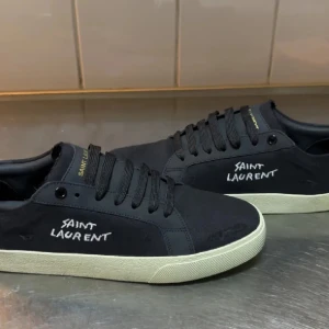 Saint Laurent sneakers - Snygga svarta sneakers från Saint Laurent med vit logga broderad på sidan och guldfärgad text på plösen. Skorna har svart snörning, vit platt sula och är tillverkade i canvas och skinn. Perfekta för dig som gillar stilrena och exklusiva detaljer.