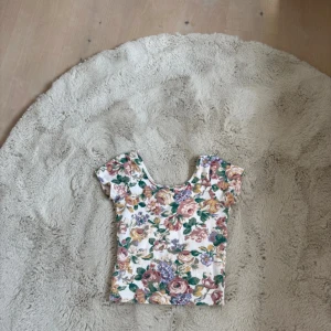 Blommig topp  - Supersöt topp från Bik Bok i storlek XS med blommigt mönster i rosa, lila, grönt och gult på vit botten. Modellen är croppad med korta ärmar och rundad urringning. Perfekt för dig som gillar romantisk och färgglad stil.