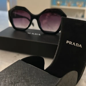 Svarta solglasögon från Prada - Snygga svarta solglasögon från Prada med cool, kantig oversized båge och mörka lila tonade glas. Kommer med originaletui och äkthetskort. Perfekt accessoar för att lyfta din stil och ge en lyxig känsla till outfiten.