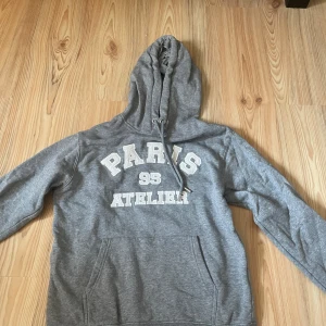 Grå hoodie med Paris-tryck  - Grå hoodie med huva och snörning, samt magficka. Tryck i vitt framtill med texten 'PARIS 95 ATELIER'. Perfekt för en chill och avslappnad stil. Mjuk insida och klassisk passform.