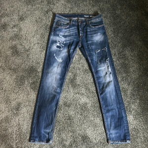 Blå distressed jeans från Reign Italia - Säljer ett par blå jeans från Reign Italia med slitna detaljer och hål på benen för en riktigt edgy look. Jeansen har klassisk femficksdesign, raka ben och är tillverkade i bomull med lite stretch för skön passform.