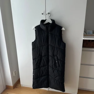 Lång svart dunväst med dragkedja - Säljer en lång svart dunväst med hög krage och praktiska fickor framtill. Västen har quiltad design, dragkedja hela vägen och tryckknappar i sidorna för extra rörelsefrihet. Perfekt att slänga över en hoodie när det är kyligt ute.